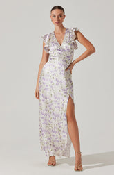A woman is wearing a long, flowy dress with a delicate floral print in shades of lavender, purple, and green on a light background. The dress features a V-neckline, ruffled short sleeves, and a high slit on one side that extends to the floor. She is also wearing nude-colored high-heeled sandals.